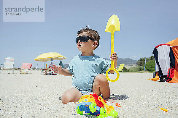 Netter Junge mit Sonnenbrille  der an einem sonnigen Tag am Strand mit Spielzeug spielt