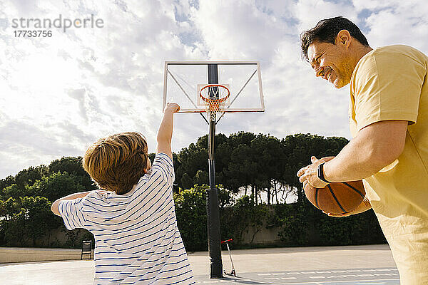 Verspielter Junge mit Vater  der an einem sonnigen Tag Basketball auf dem Sportplatz hält