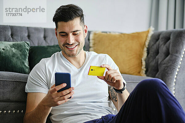 Lächelnder Mann mit Kreditkarte beim Online-Einkauf per Smartphone zu Hause