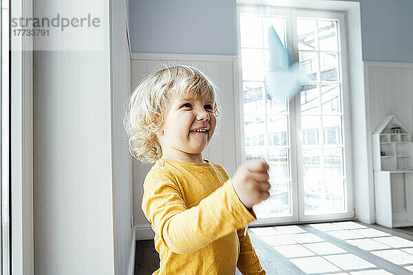 Cute boy playing with origami bird standing at home