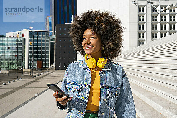 Lächelnde  schöne junge Frau mit kabellosen Kopfhörern  die mit ihrem Mobiltelefon an der Treppe steht