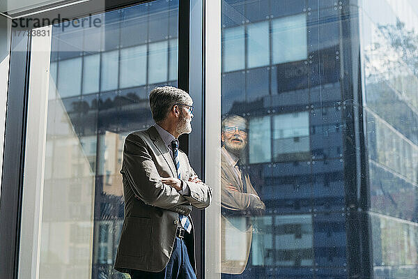 Geschäftsmann mit verschränkten Armen blickt durch Fenster im Büro