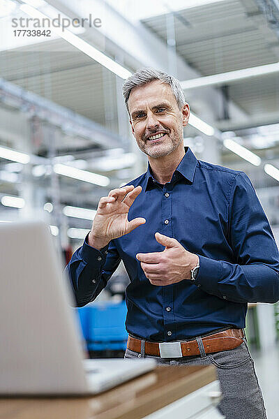 Reifer Geschäftsmann gestikuliert während eines Videoanrufs auf einem Laptop in der Fabrik