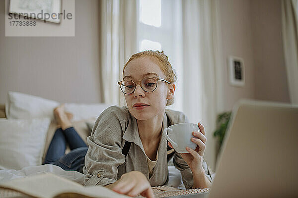 Frau hält Kaffeetasse in der Hand und liest ein Buch  das zu Hause auf dem Bett liegt