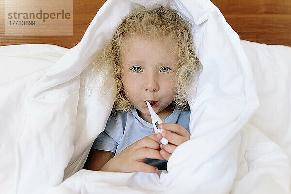 Krankes Mädchen in Decke gehüllt mit digitalem Thermometer zu Hause