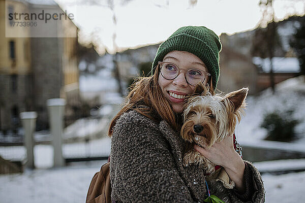 Lächelnde Frau mit Strickmütze umarmt Yorkshire Terrier