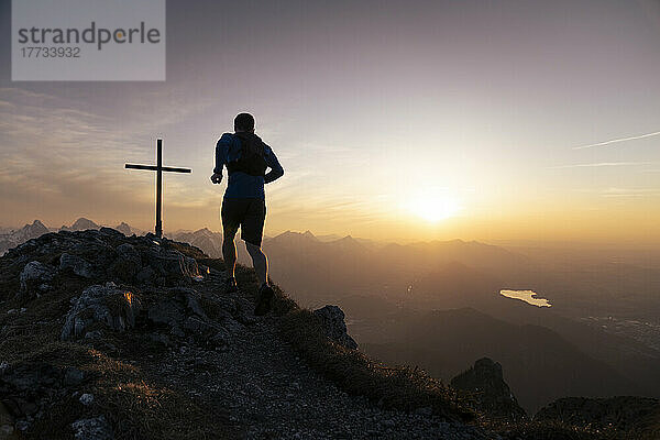 Wanderer läuft auf dem Sauling-Berggipfel mit Gipfelkreuz bei Sonnenuntergang