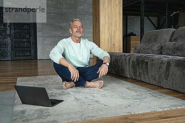 Mann mit geschlossenen Augen macht Yoga mit Laptop auf Teppich zu Hause