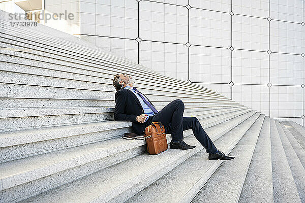Reifer Geschäftsmann sitzt neben seiner Tasche und entspannt sich auf Stufen