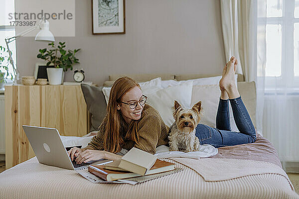 Lächelnde Frau liegt mit Laptop und schaut auf den Hund  der zu Hause im Bett sitzt