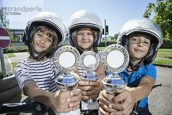 Smiling friends wearing helmet showing trophies on sunny day