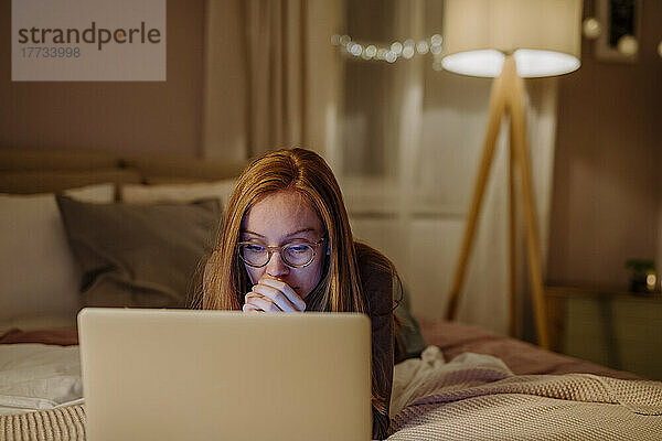 Frau mit Brille und Laptop liegt zu Hause im Bett