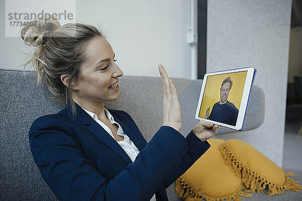 Eine Geschäftsfrau gestikuliert bei einem Videoanruf über einen Tablet-PC und sitzt auf dem Sofa im Büro