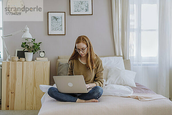 Frau sitzt mit gekreuzten Beinen zu Hause im Bett und benutzt Laptop
