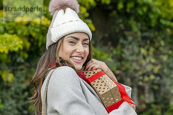 Schöne glückliche Frau mit Strickmütze umarmt Weihnachtsgeschenk im Park