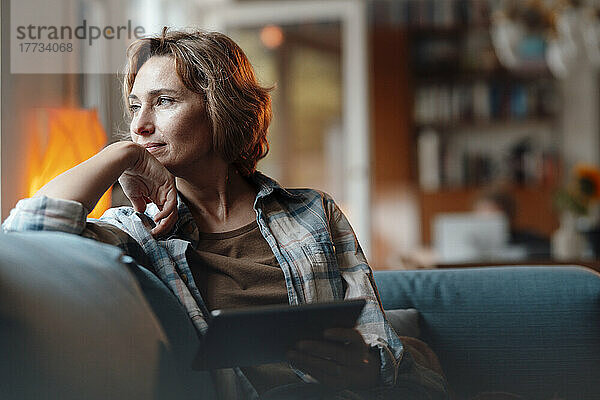Thoughtful woman with tablet PC sitting on sofa at home
