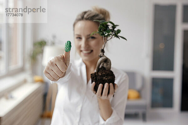 Glückliche Geschäftsfrau hält Pflanze und zeigt grünen Daumen im Büro