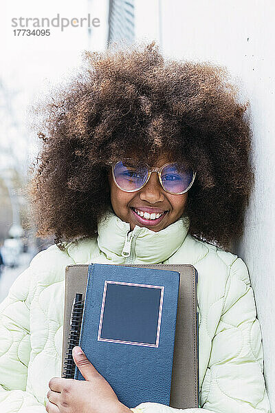 Lächelndes Afro-Schulmädchen mit Brille und Büchern an der Wand