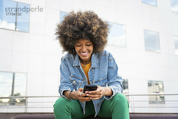 Fröhliche Afro-Frau schreibt Textnachrichten per Smartphone und sitzt auf einer Bank