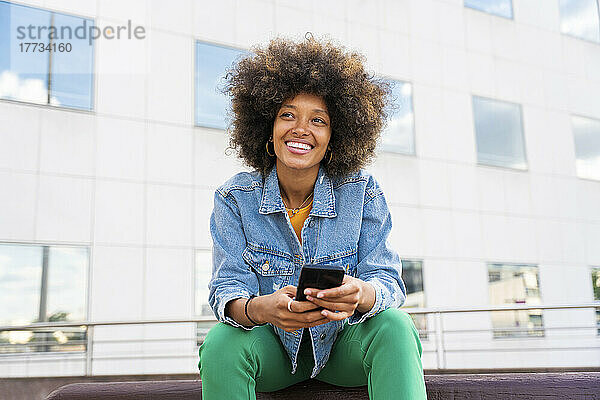 Schöne glückliche Frau mit Afro-Frisur  die ihr Mobiltelefon auf einer Bank hält