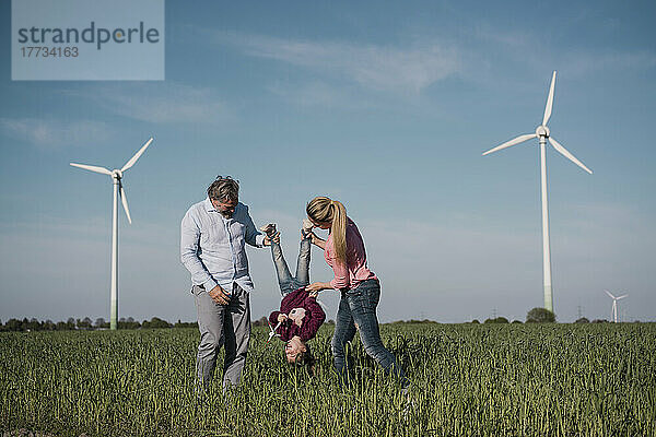 Verspielte Eltern tragen ihre Tochter kopfüber mit Windkraftanlagen im Hintergrund im Windpark