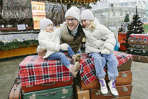 Lächelnde Mädchen sitzen auf Koffern des Vaters auf dem Weihnachtsmarkt