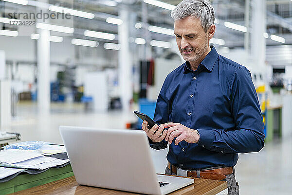 Reifer Geschäftsmann nutzt Laptop und Smartphone in der Fabrik
