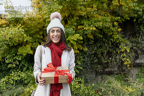 Fröhliche junge Frau mit Strickmütze steht mit Weihnachtsgeschenk im Park
