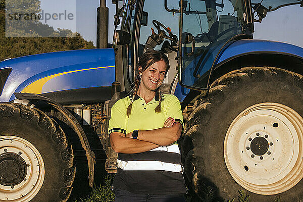 Lächelnde Bäuerin steht mit verschränkten Armen vor dem Traktor