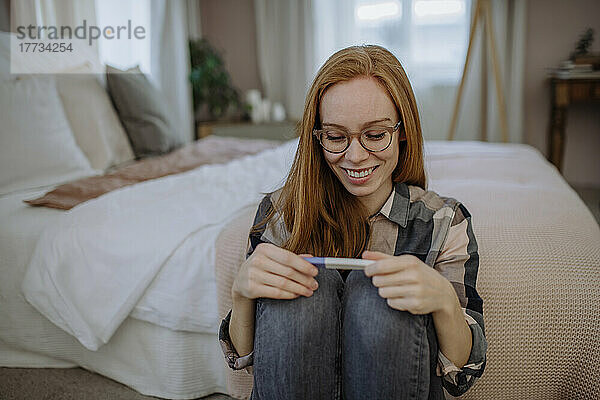 Glückliche Frau mit Schwangerschaftstestset sitzt zu Hause vor dem Bett
