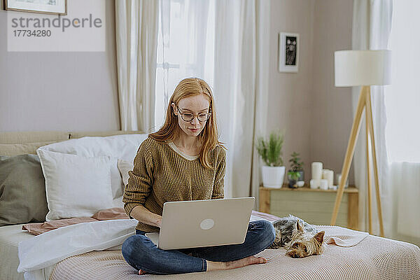 Frau sitzt mit gekreuzten Beinen und Hund mit Laptop im heimischen Bett