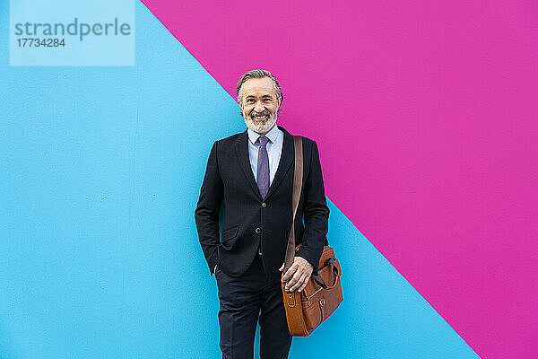 Glücklicher reifer Geschäftsmann mit Tasche  der vor einer rosa und blauen Wand steht