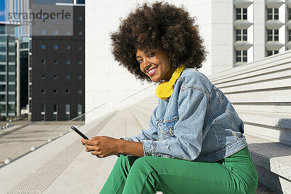 Lächelnde schöne Afro-Frau mit kabellosen Kopfhörern sitzt mit Mobiltelefon auf Stufen