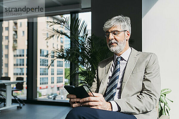 Lächelnder Geschäftsmann  der Textnachrichten über sein Mobiltelefon im Büro sendet