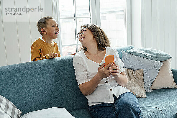 Fröhlicher Junge spricht mit seiner Mutter  die zu Hause auf dem Sofa sitzt und ihr Mobiltelefon in der Hand hält