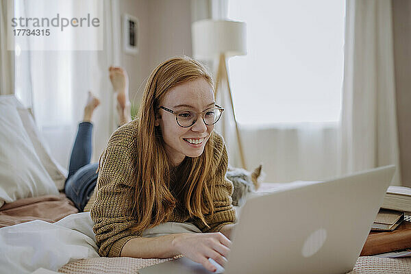 Lächelnde Frau mit Brille und Laptop  die zu Hause im Bett liegt