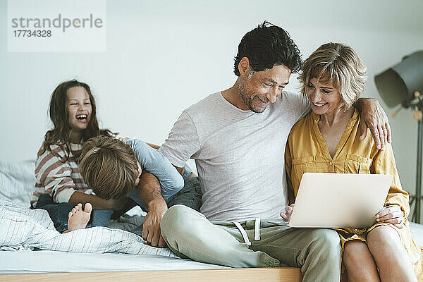 Glücklicher Mann und Frau mit Laptop sitzen neben Tochter und Sohn und spielen zu Hause im Bett