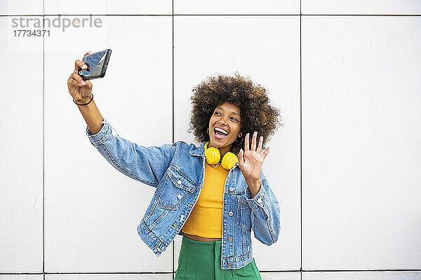 Glückliche Afro-Frau winkt bei Videoanruf über Smartphone vor der Wand mit der Hand