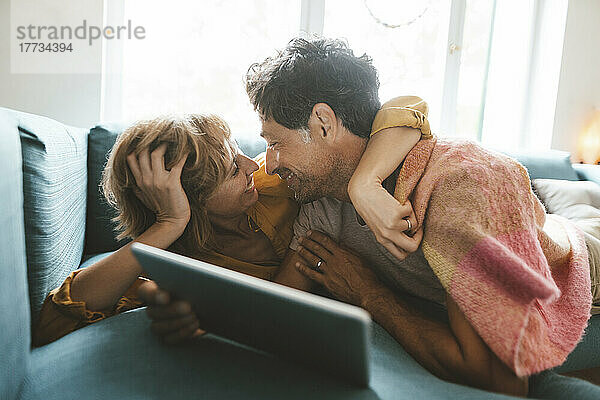 Lächelnder Mann mit Tablet-PC umarmt Frau  die zu Hause auf dem Sofa liegt