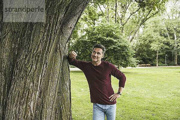 Lächelnder reifer Mann mit der Hand auf der Hüfte und Blick auf den Baum im Park