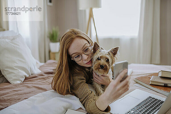 Lächelnde Frau macht ein Selfie mit ihrem Hund über ihr Smartphone  das zu Hause im Bett liegt