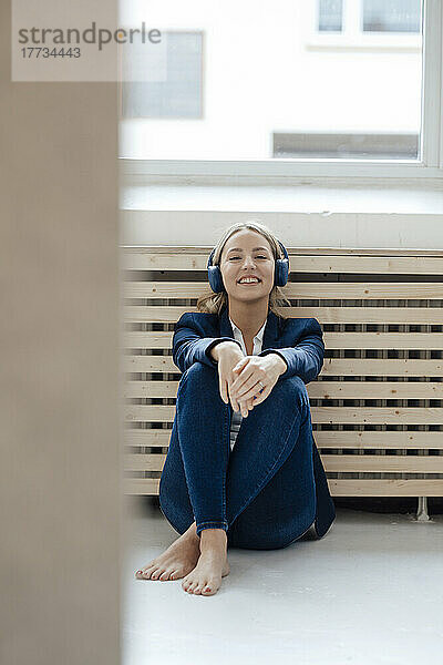 Lächelnde Geschäftsfrau  die im Büro Musik über drahtlose Kopfhörer hört und sich an den Heizkörper lehnt