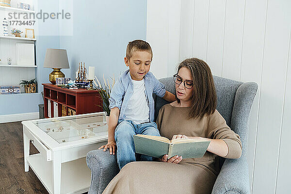 Mutter liest ihrem Sohn ein Buch vor  der zu Hause auf einem Sessel sitzt