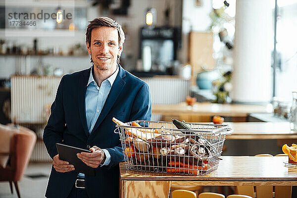 Lächelnder Geschäftsmann mit Tablet-PC neben dem Korb  der sich im Café auf den Tisch lehnt