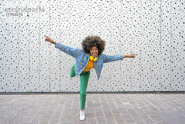 Glückliche Afro-Frau mit ausgestreckten Armen  die auf einem Bein vor der Wand steht