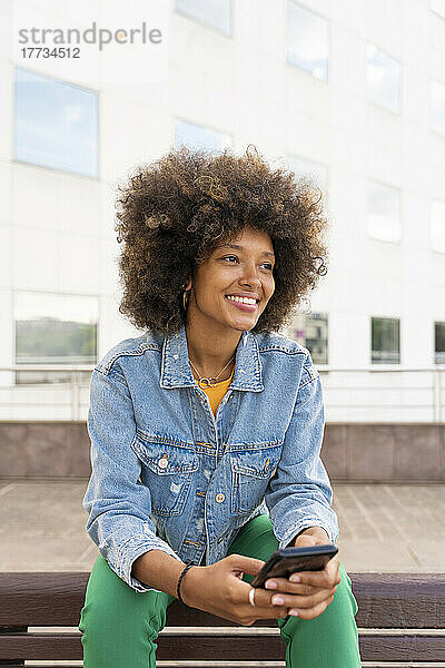 Lächelnde Frau mit Afro-Frisur hält Smartphone auf Bank sitzend