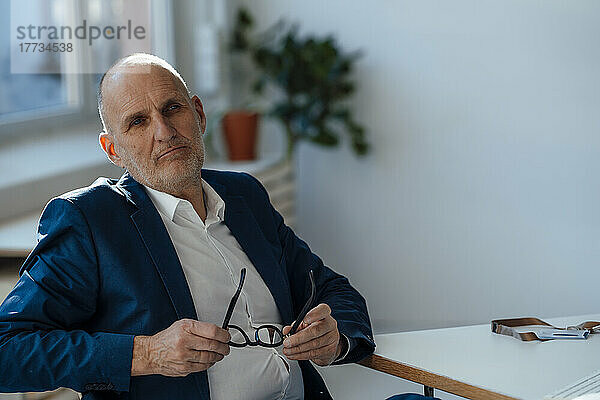 Businessman holding eyeglasses sitting by desk in office