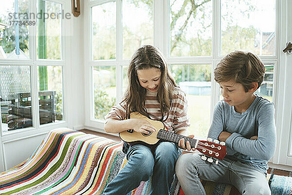 Lächelndes Mädchen  das Gitarre spielt und mit seinem Bruder zu Hause sitzt