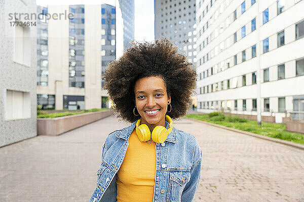 Lächelnde schöne Afro-Frau mit kabellosen Kopfhörern steht auf dem Fußweg
