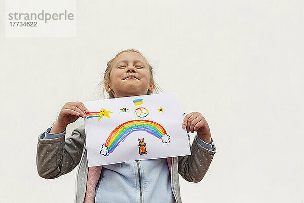 Mädchen mit geschlossenen Augen zeigt Regenbogen und Friedenssymbol auf Papier vor weißem Hintergrund